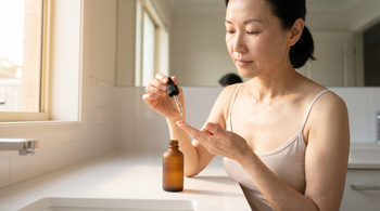 51-year-old East Asian Australian woman with fair skin applying a serum to her fingertips at a white bathroom vanity in warm morning light