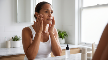 47-year-old Pacific Islander Australian woman with brown skin applying a lightweight serum to her cheek in a bright bathroom with natural morning light