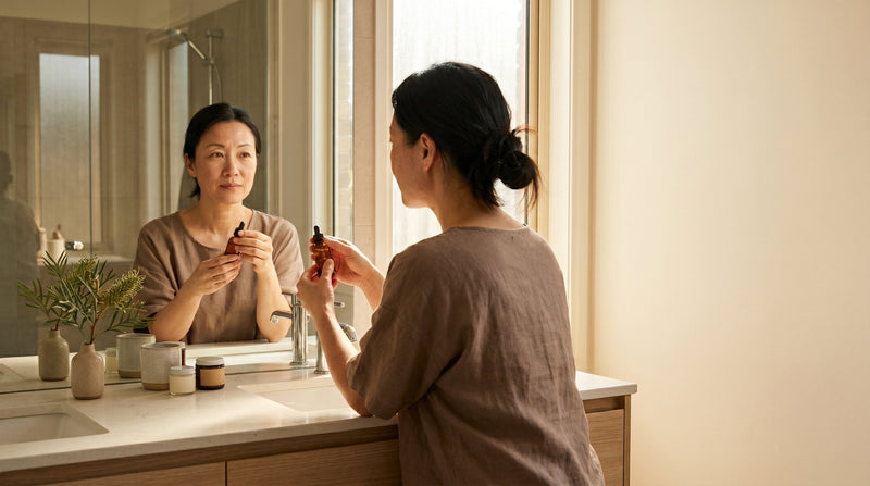 Woman examining her skin in a bathroom mirror while holding a skincare treatment bottle in morning light
