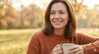 A woman in her 40s with natural, glowing skin smiling softly in a sunlit autumn park, holding a warm ceramic mug to illustrate the shift to a comforting skincare ritual.