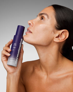 Woman with glowing skin looking upward, holding a purple Dermalogica Phyto Nature e² bottle near her chin. A small amount of product is applied to her cheek, set against a soft grey gradient background.